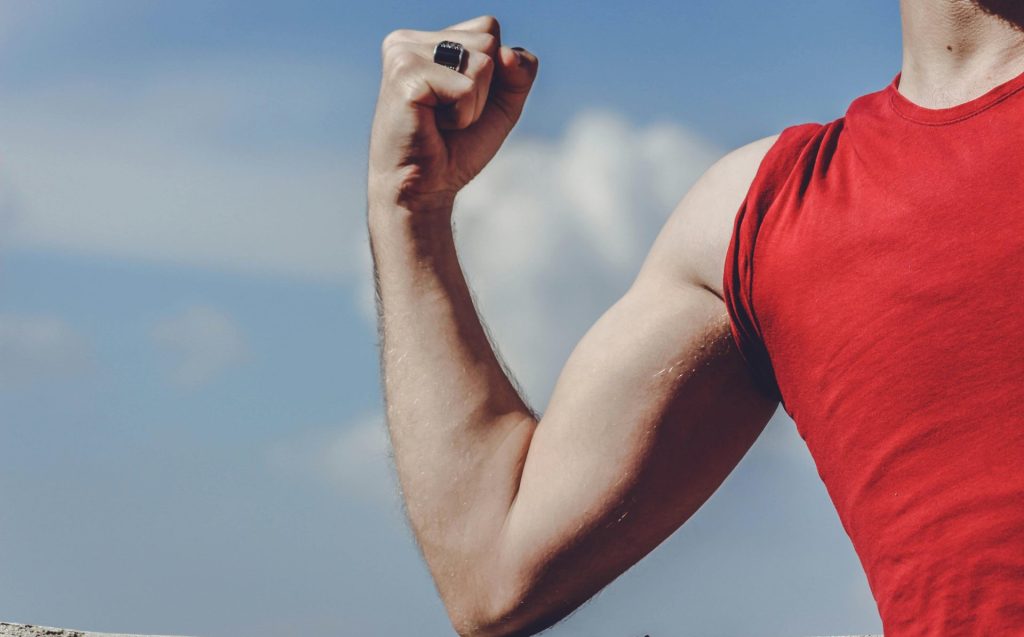 Nahaufnahme eines gebeugten Arms in einem roten Hemd vor einem klaren blauen Himmel, der Stärke und Ermächtigung symbolisiert.