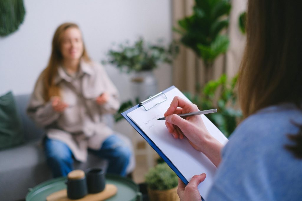 Nicht erkennbare professionelle Psychologin, die während einer Psychotherapiesitzung im hellen Büro auf der Zwischenablage schreibt, während sie vor dem Klienten auf unscharfem Hintergrund sitzt